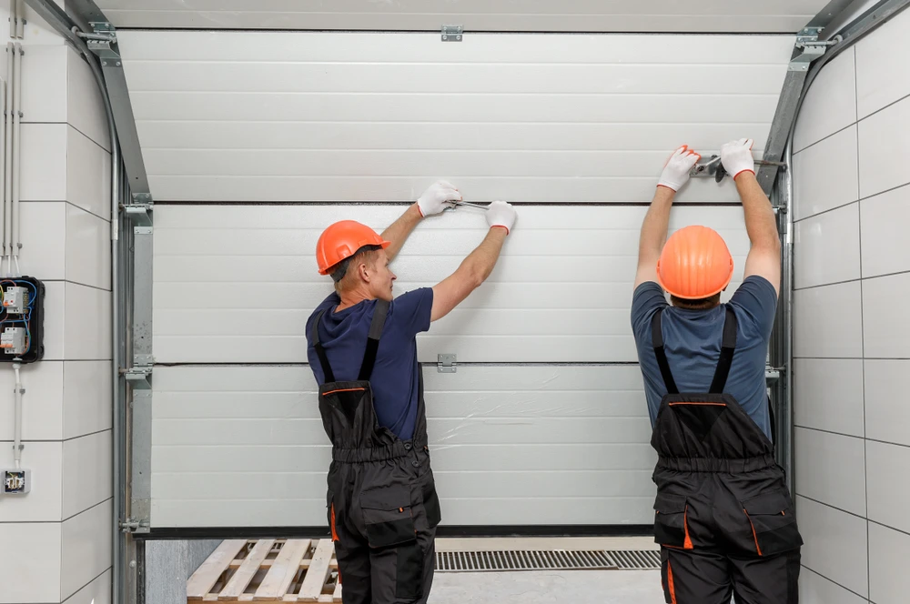 Deux techniciens en combinaisons de travail et casques orange installent une porte de garage sectionnelle blanche dans un garage r&eacute;sidentiel