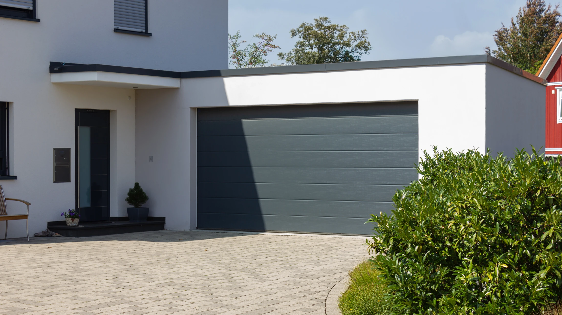 Photographie en gros plan d'une main masculine actionnant une télécommande moderne de motorisation de porte de garage, avec en arrière-plan une porte de garage sectionnelle fermée en aluminium gris ou blanc. La scène se déroule dans le garage, de jour, avec l'espace intérieur du garage partiellement visible. L'éclairage doit être naturel et professionnel, mettant en avant le geste de commande et la technologie de la télécommande.